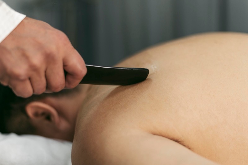 En hand för ett mörkt skrapverktyg över ryggen på en person som ligger på en massagebänk